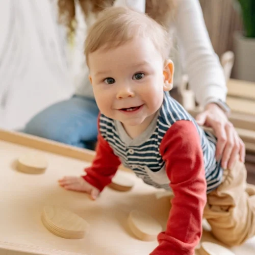 Un bébé souriant avec des cheveux blonds, portant un t-shirt à rayures bleues et blanches avec des manches rouges, qui joue sur une table en bois avec des éléments en bois
