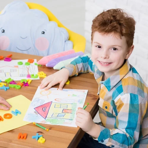 Un garçon avec des cheveux bouclés roux, souriant, assis à une table, en train de colorier une maison dessinée sur une feuille. Il y a des jouets en plastique colorés et un coussin en forme de nuage dans l'arrière-plan.