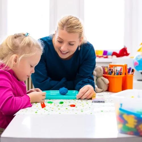 Une femme et une jeune fille jouent avec de la pâte à modeler sur une table dans une pièce lumineuse.