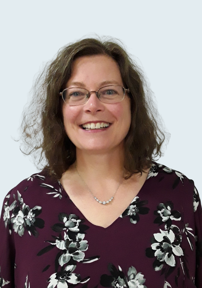 Une femme souriante avec des lunettes, des cheveux bouclés bruns, portant un chemisier violet avec des fleurs blanches et un collier