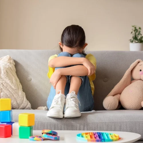 Une enfant assise sur un canapé, les bras autour de ses jambes, visage caché, entourée de jouets