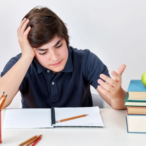 Un jeune garçon semble confus ou frustré devant un cahier ouvert, avec des livres empilés à côté. Il tient une main à la tête et l'autre main tendue vers le cahier.