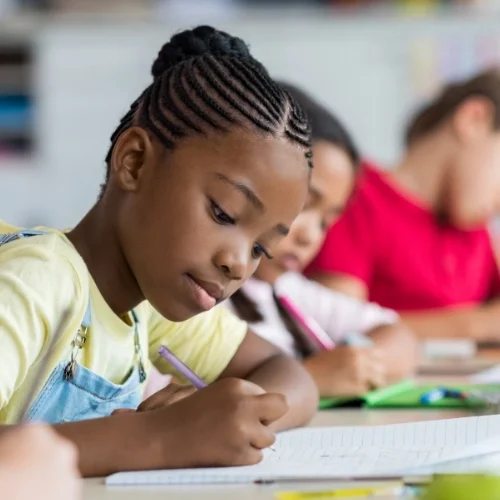 Une élève africaine écrit dans un cahier en classe, avec d'autres élèves en arrière-plan.