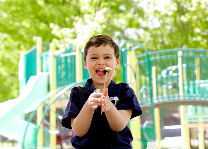 Un garçon souriant tenant une fleur, dans un parc pour enfants avec des structures de jeu en arrière-plan.