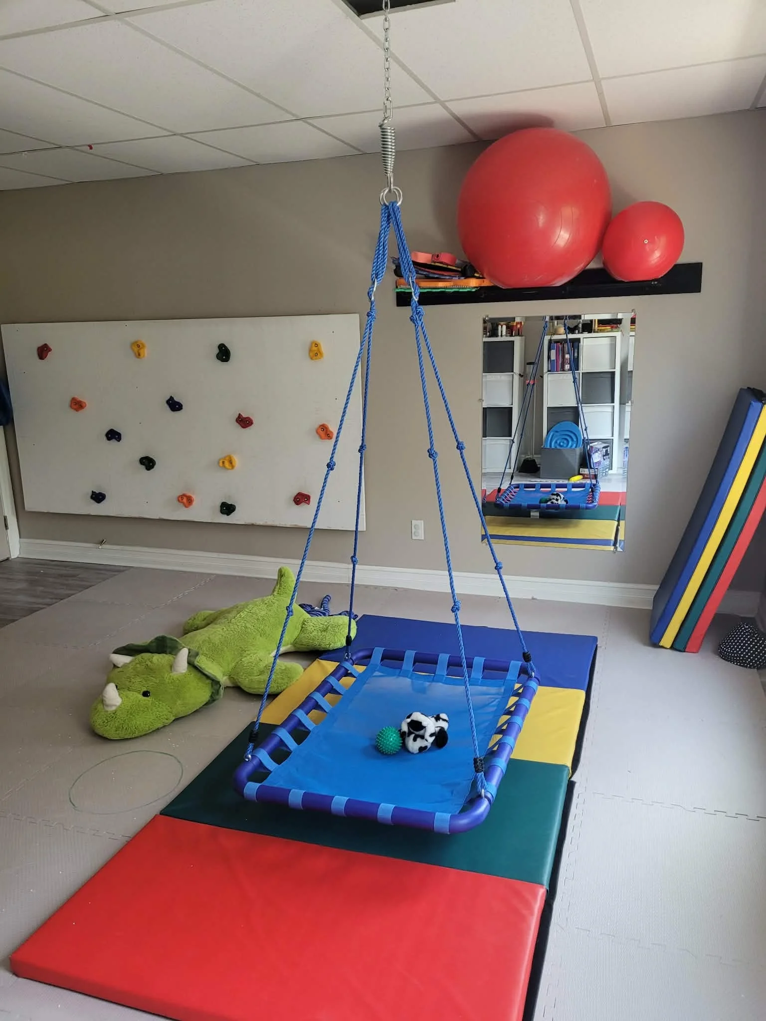 Une salle de jeux pour enfants avec une balançoire en filet suspendue au plafond, un tapis de sol coloré, un gros doudou crocodile vert, des balles et une aire de jeux avec des ballons et des étagères remplies de jouets et de livres.