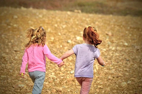 Due bambine che corrono e si tengono per mano in un parco autunnale con foglie cadute sul terreno.