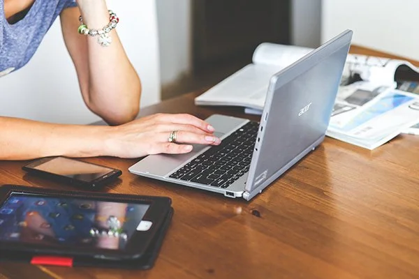 Una persona che lavora su un laptop su un tavolo di legno, con uno smartphone e un tablet accanto, e alcune riviste o documenti aperti.