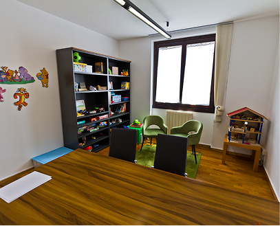 Stanza per bambini con scaffale pieno di giochi, due sedie verdi, un tavolo di legno e una piccola casa delle bambole.