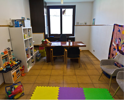 Una stanza con una scrivania, tre sedie, una libreria con giocattoli e libri, una sedia con uno sfondo colorato e un pavimento di piastrelle.