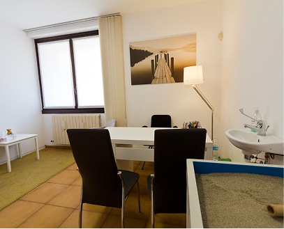 Stanza con tavolo bianco, quattro sedie nere, lampada da tavolo, quadro con scena di un molo e un lago, finestra con tende chiare, lavandino in angolo, pavimento di piastrelle marroni e soffitto bianco.