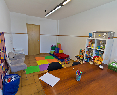 Sala giochi per bambini con tappeti colorati, giochi impilabili, cartelle, cassetti con giocattoli e tavolo con penne colorate.
