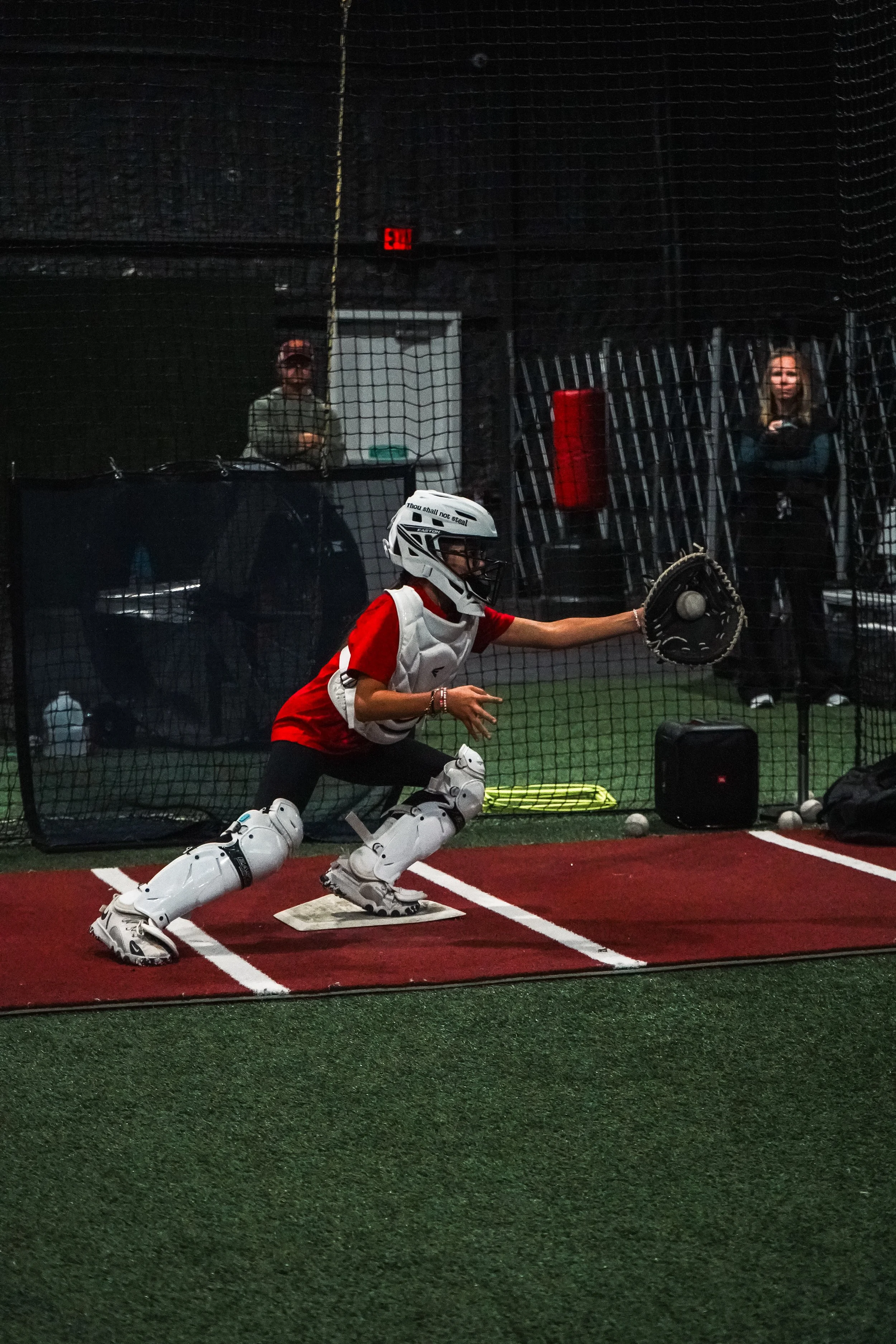 Youth baseball catcher in a red uniform and protective gear, crouching behind home plate on a sports field, with a baseball in hand, preparing to throw it. In the background, a woman standing with arms crossed and another person wearing a baseball cap are visible, separated by a net.