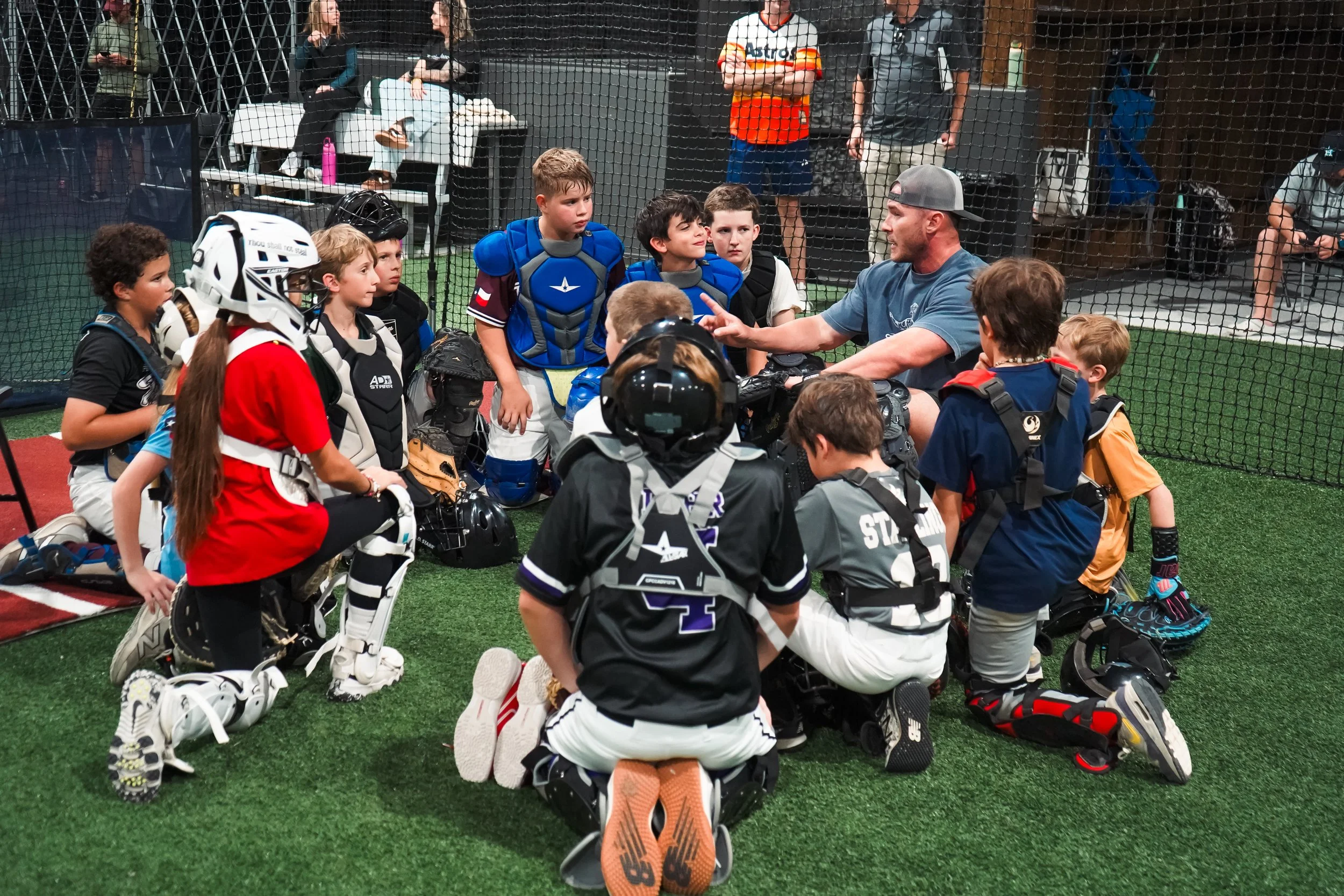 Youth baseball players and coaches having a discussion inside a batting cage, with some players wearing catcher's gear, while others are kneeling or sitting on the turf.