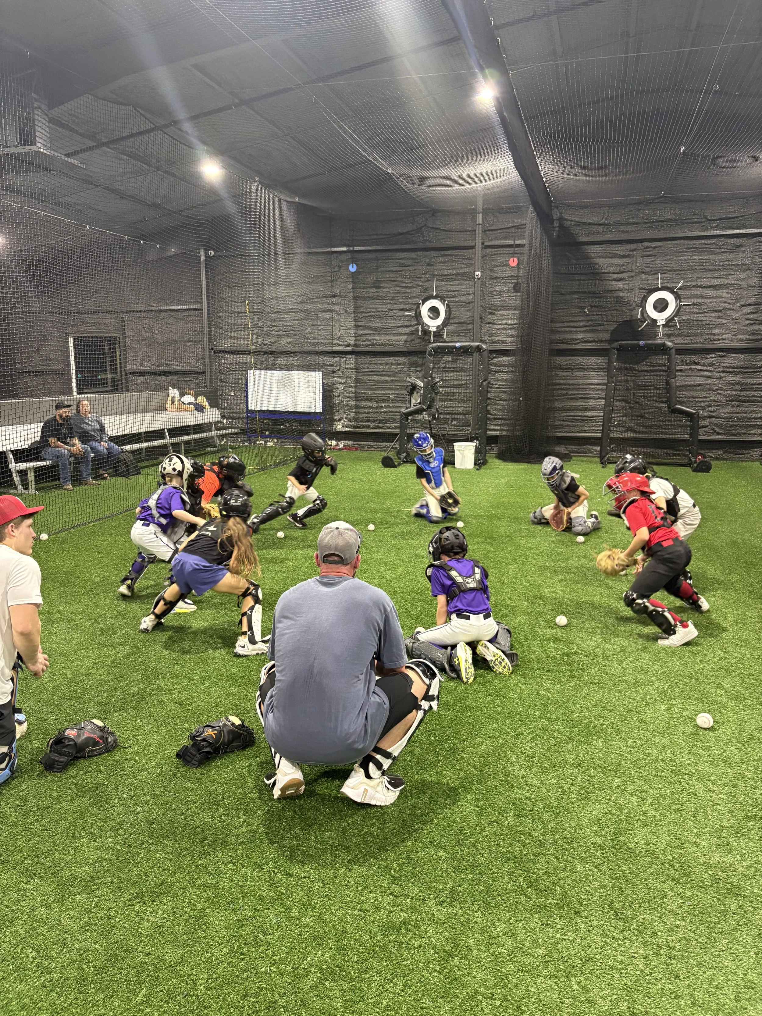 Young children playing baseball indoors on artificial turf, wearing helmets and gloves, with some adults watching and sitting on benches behind a protective net.