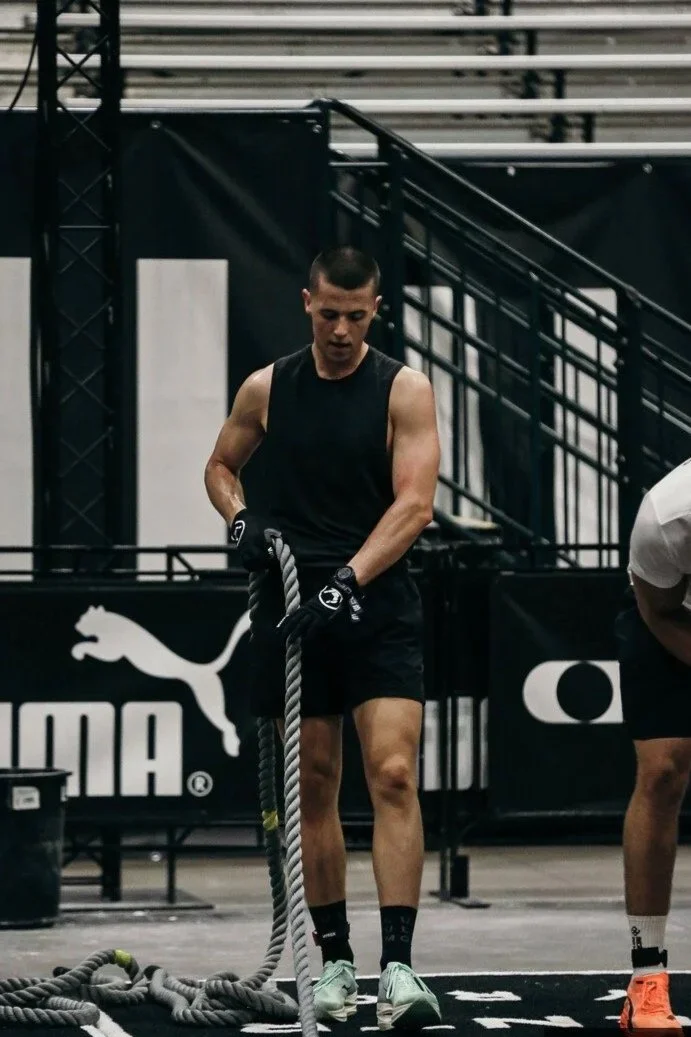 A young man in a black sleeveless shirt and shorts is doing battle rope exercises in a gym with black and white Puma branding and metal stairs in the background.