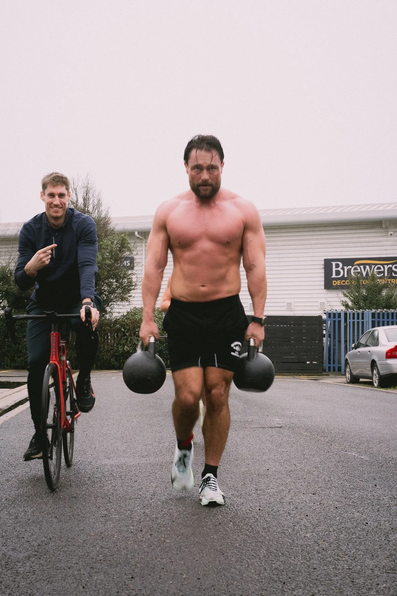 A shirtless man running with kettlebells in each hand, and a smiling man riding a bicycle nearby, on a gray overcast day in a parking lot.