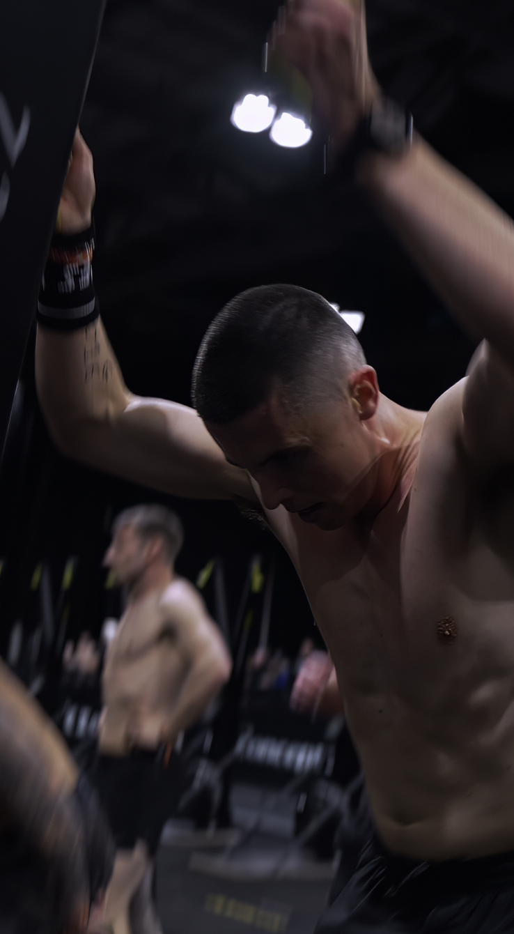 A shirtless man with a short haircut looking down, holding a bottle, in a dimly lit gym with other people working out in the background.