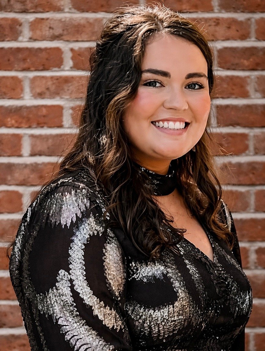 A young woman with long brown wavy hair smiling at the camera, wearing a black and silver sequined dress, standing in front of a brick wall.