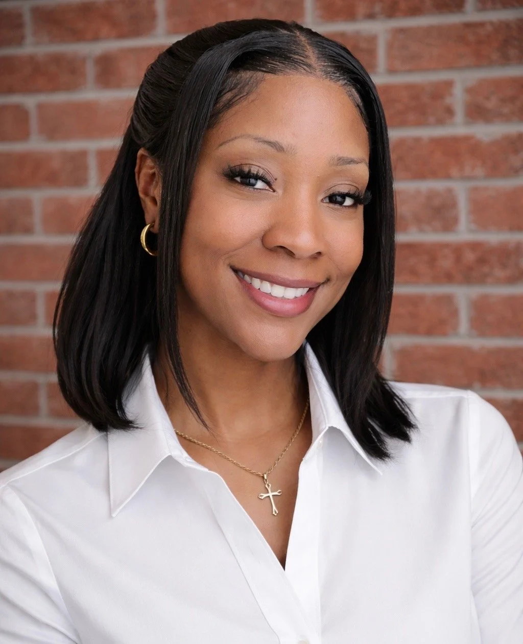 A woman with shoulder-length black hair, wearing a white collared shirt, gold hoop earrings, a gold necklace with a cross pendant, and smiling against a brick wall background.