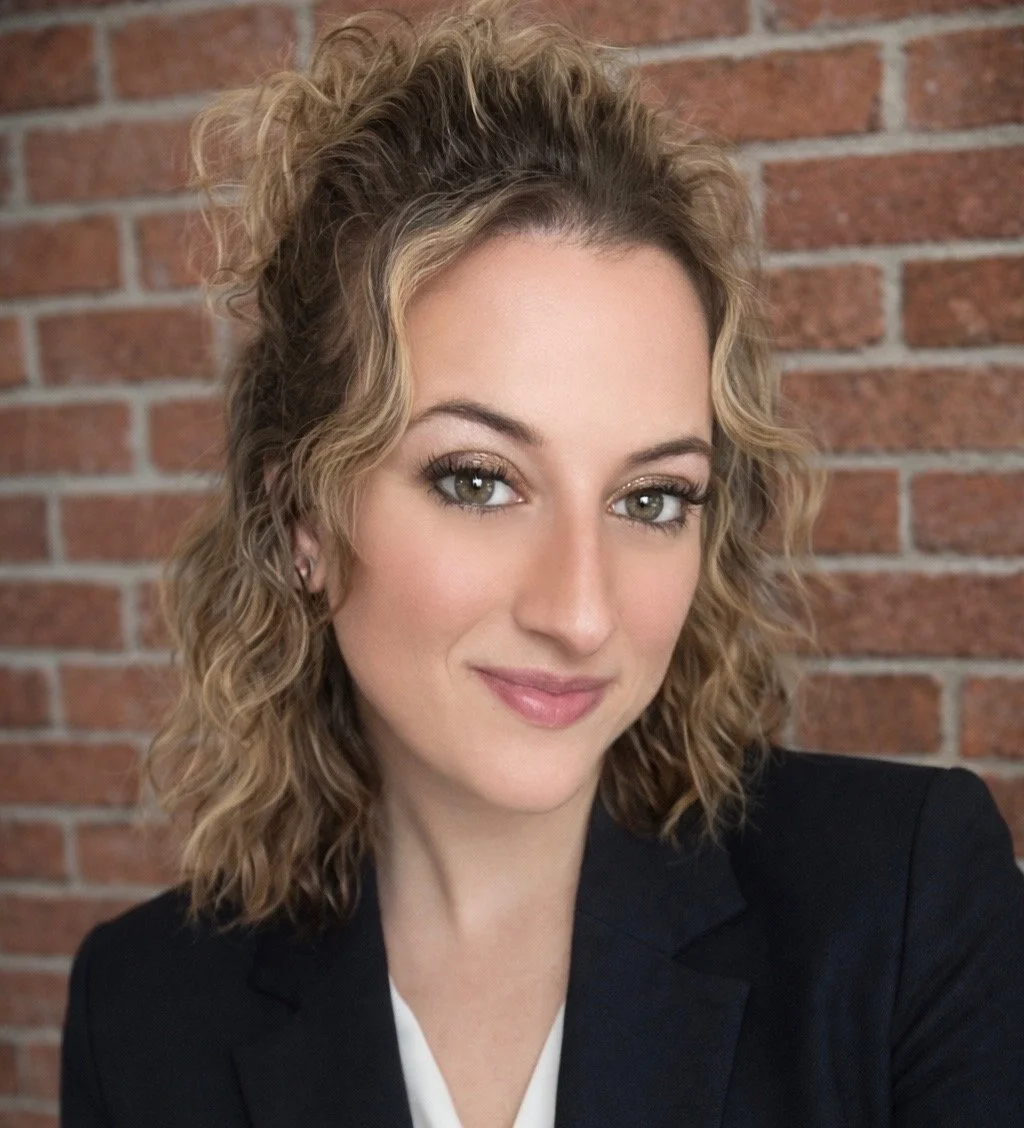 A young woman with curly blonde hair, wearing a black blazer, smiling against a brick wall background.