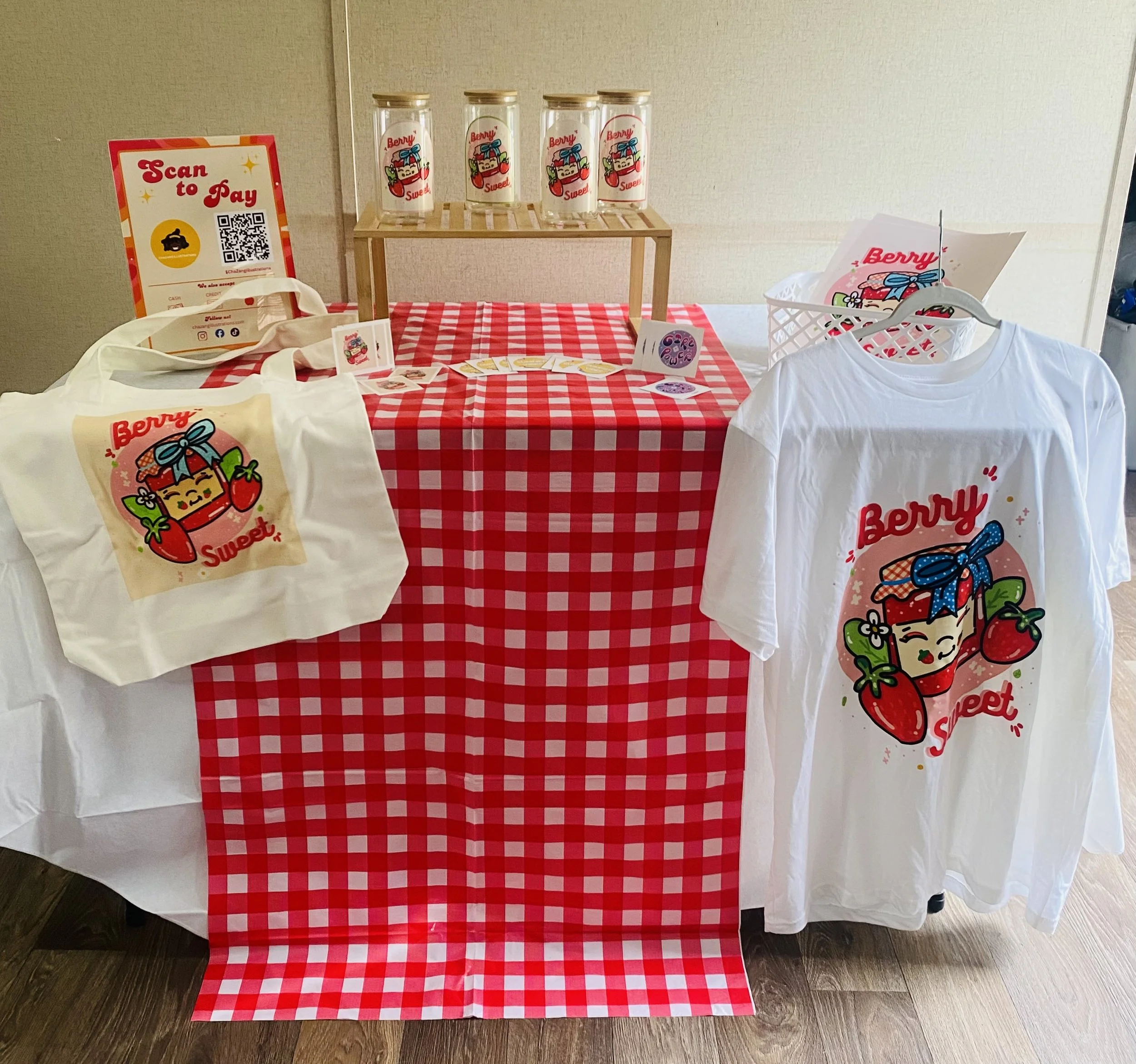 Display table with red and white checkered tablecloth, promotional items, and jars with cartoon characters and strawberries, related to Berry Sweet branding.