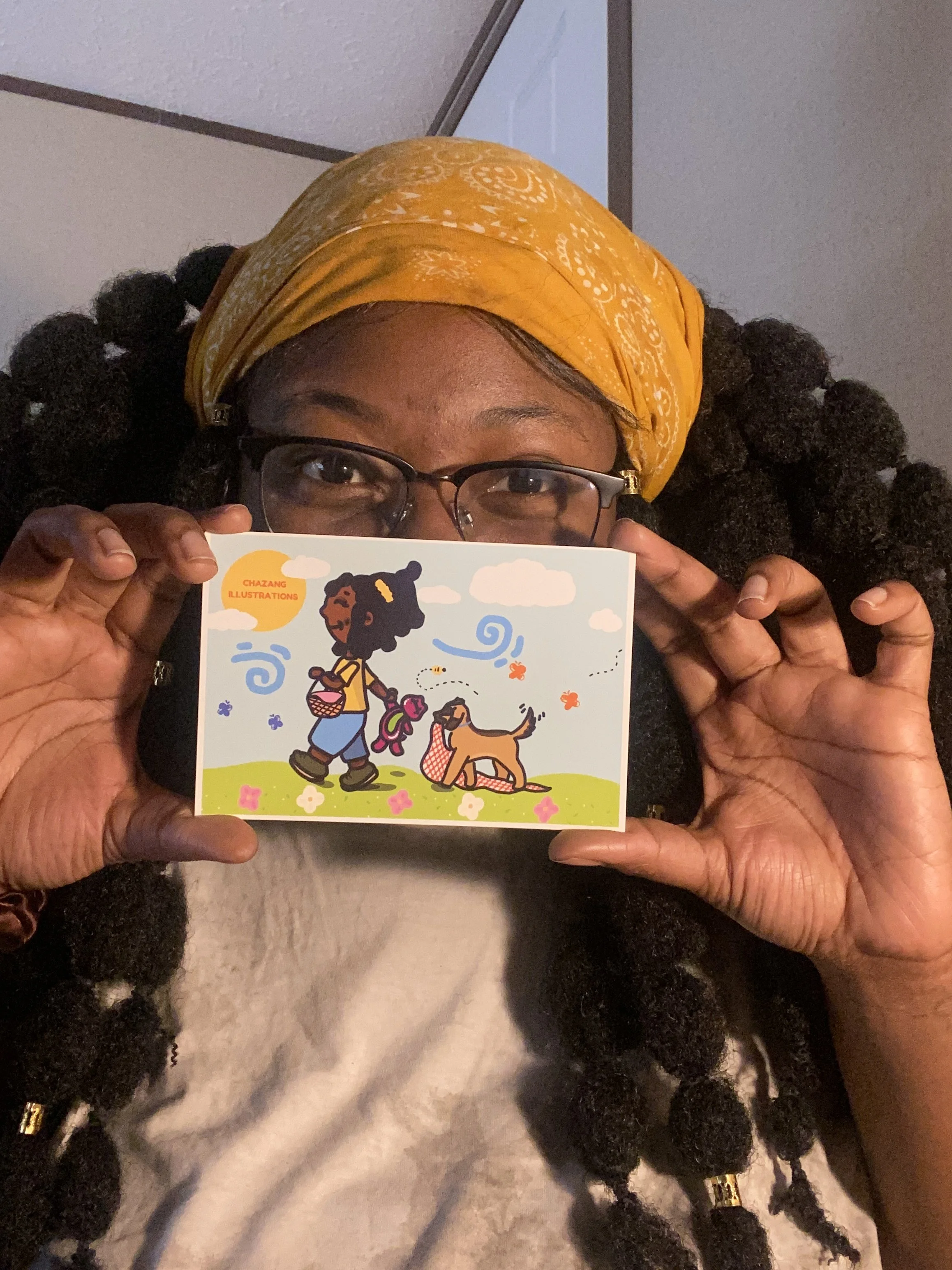 A person with glasses and braids wearing a yellow headscarf holding a colorful illustrated card with a girl and a dog, titled 'ChaZang Illustrations'.