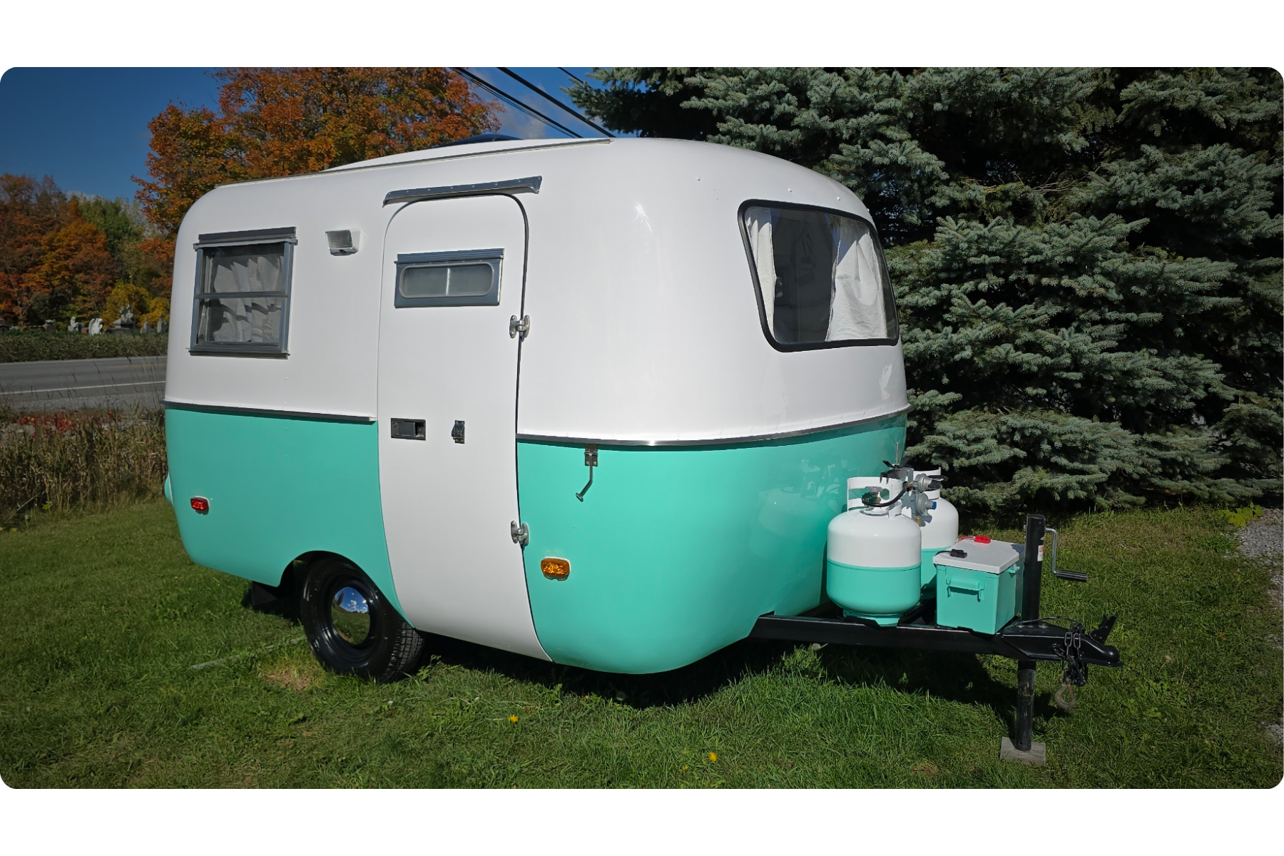 Caravane vintage blanche et verte avec fenêtres, stationnée sur une pelouse avec un arbre et un ciel bleu en arrière-plan.