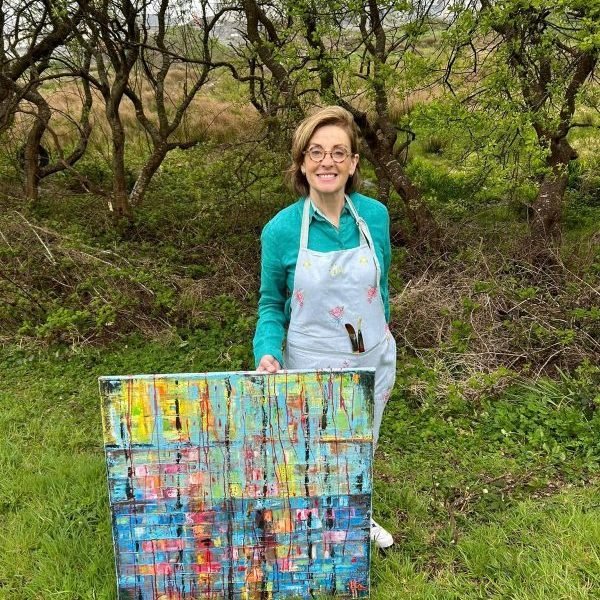 A woman wearing glasses, a teal sweatshirt, and an apron, standing outdoors in a grassy area with trees in the background. She is holding a colorful abstract painting.