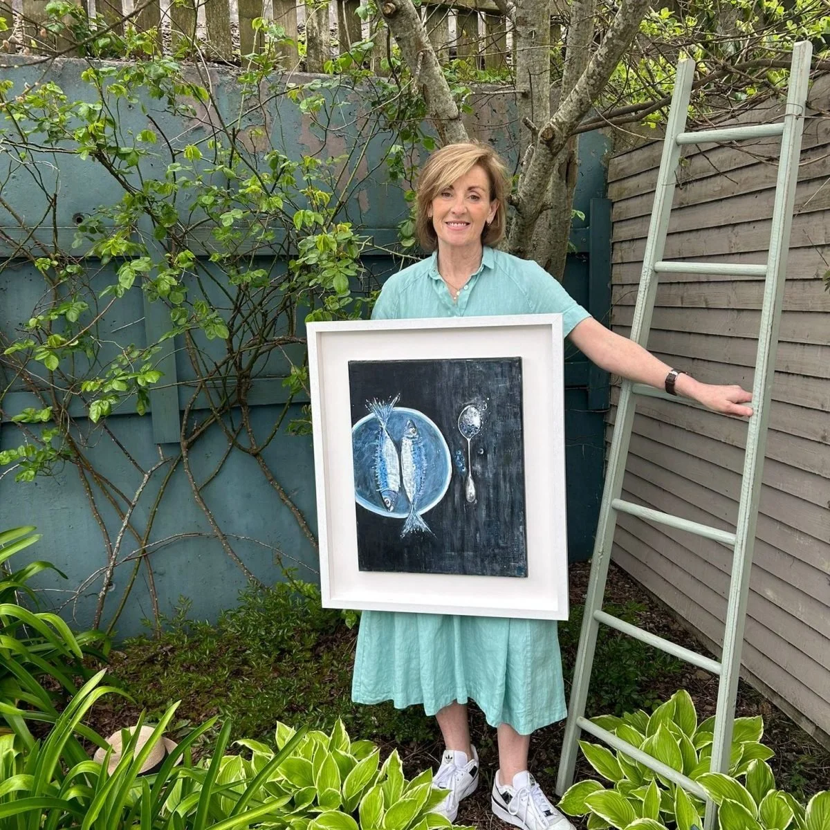 A woman in a light green dress holding a framed painting of two fish and a spoon, standing outdoors next to a ladder and surrounded by greenery.