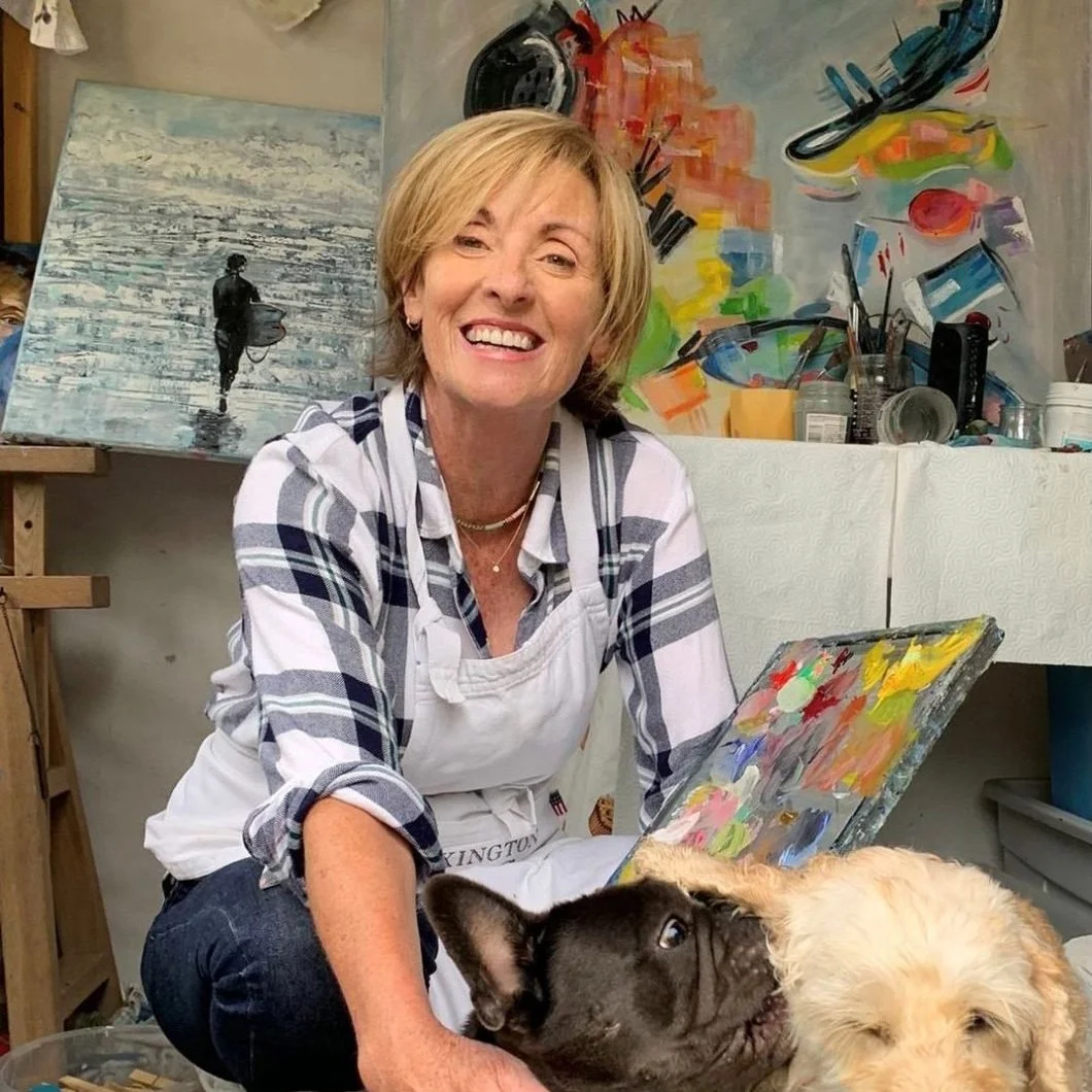A woman with short blonde hair smiling while sitting in an art studio, accompanied by two dogs, one black and one light-colored. Behind her, there are colorful paintings and art supplies.