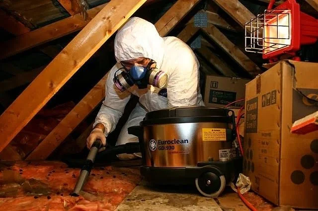 Person wearing protective gear uses a vacuum in an attic filled with insulation and boxes.