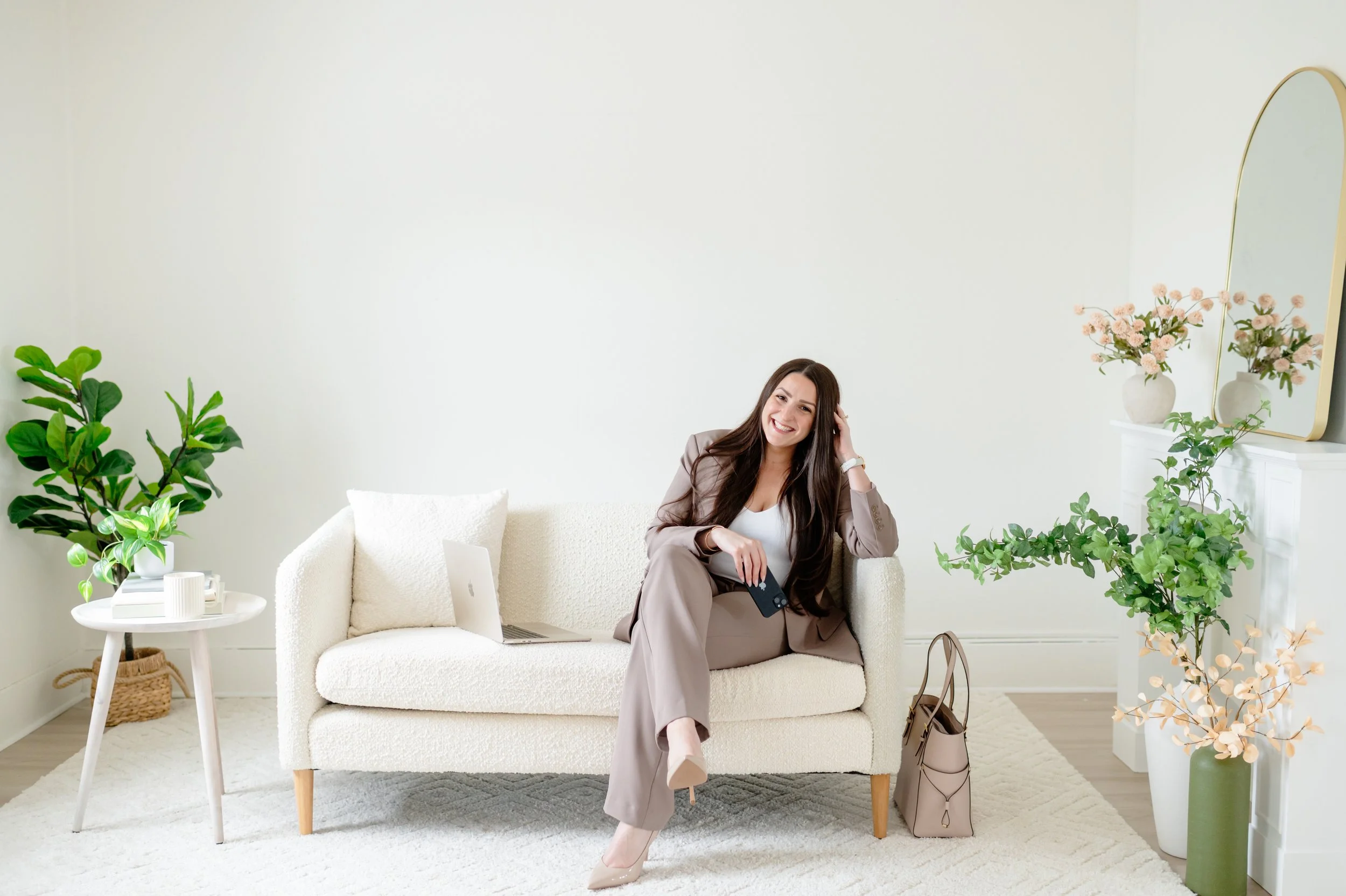 A woman sitting on a cream-colored sofa in a bright room, smiling, holding a remote, with a laptop beside her. The room has green plants, a white side table with books and a cup, and a white fireplace with a mirror and pink flowers.