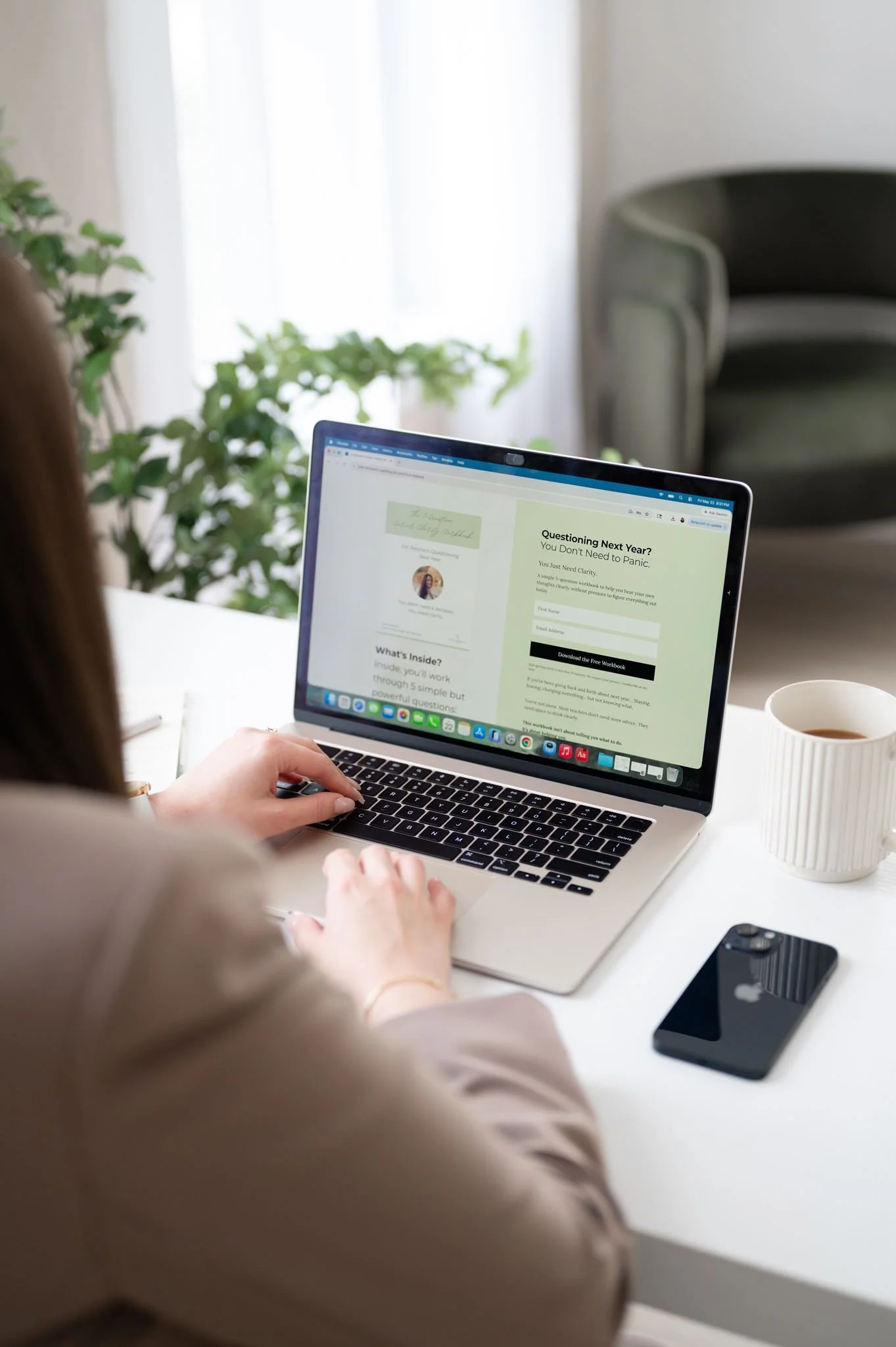 Person using a MacBook laptop at a white desk with a cup of coffee, a smartphone, and a potted plant in a bright, modern room.