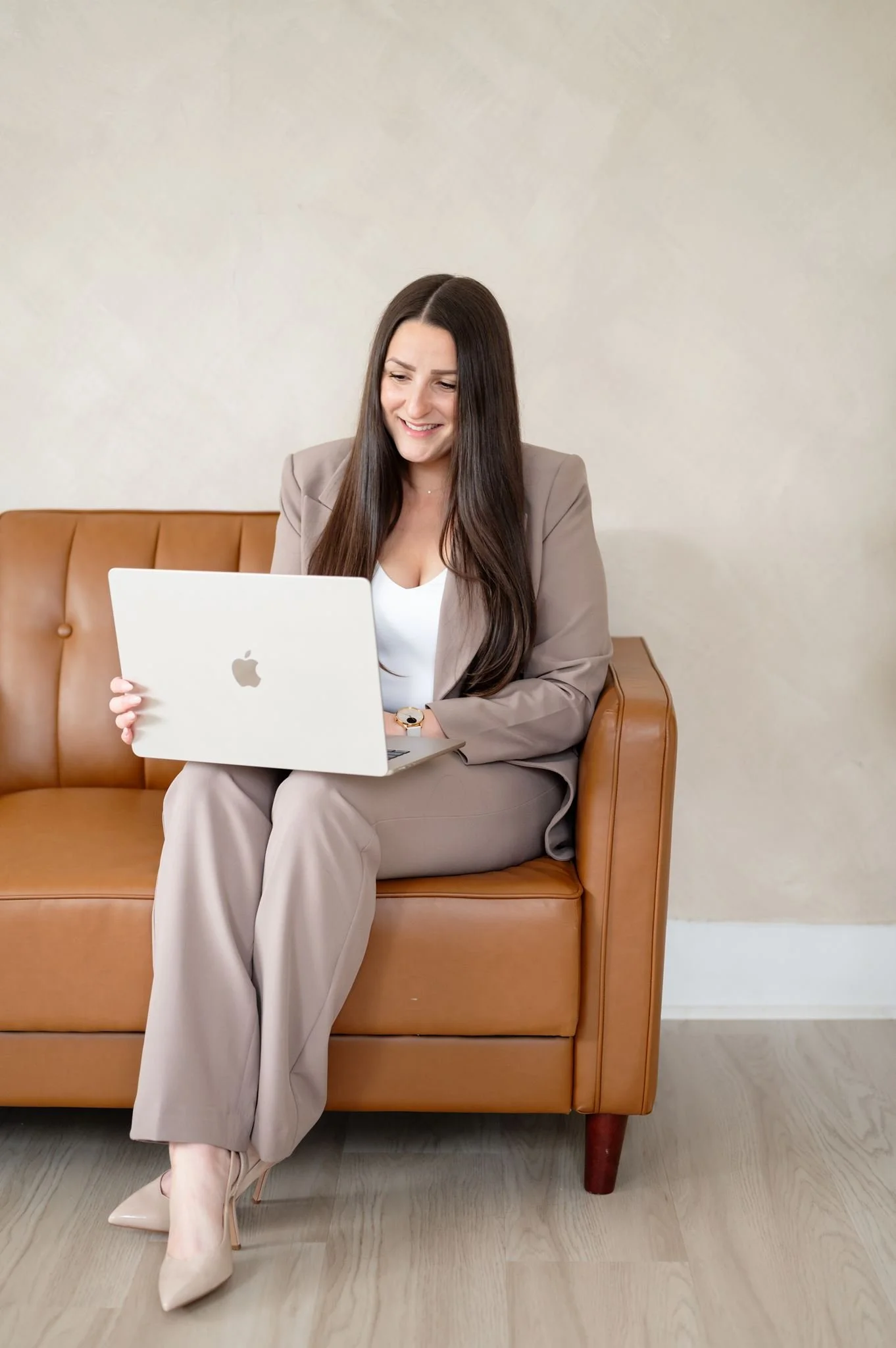 A woman with long dark hair sitting on a tan leather sofa, holding a silver MacBook laptop and smiling. She is dressed in beige pants, a beige blazer, and a white top, wearing beige high heels, with a light-colored background and wooden flooring.