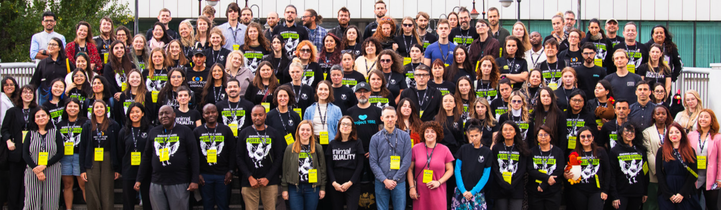 A large group of diverse people gathered outside in front of a white modern building, posing for a group photo at the Open Wing Alliance Global Summit in 2025. Many are wearing black shirts and yellow lanyards with animal advocacy messages.