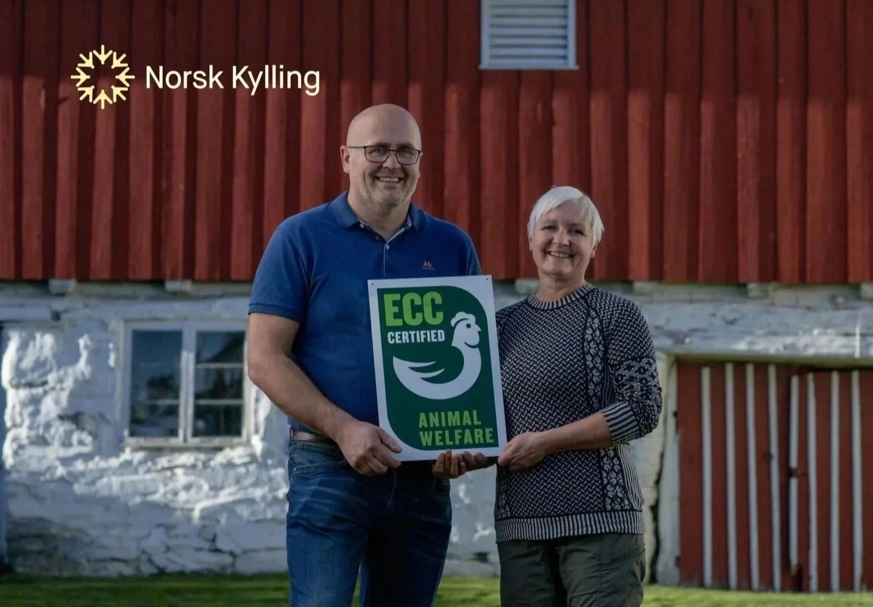 Two people, a man and a woman, standing outside in front of a red wooden building. The man is holding a sign that reads "ECC Certified Animal Welfare" with a logo featuring a stylized bird or animal. They are smiling at the camera.