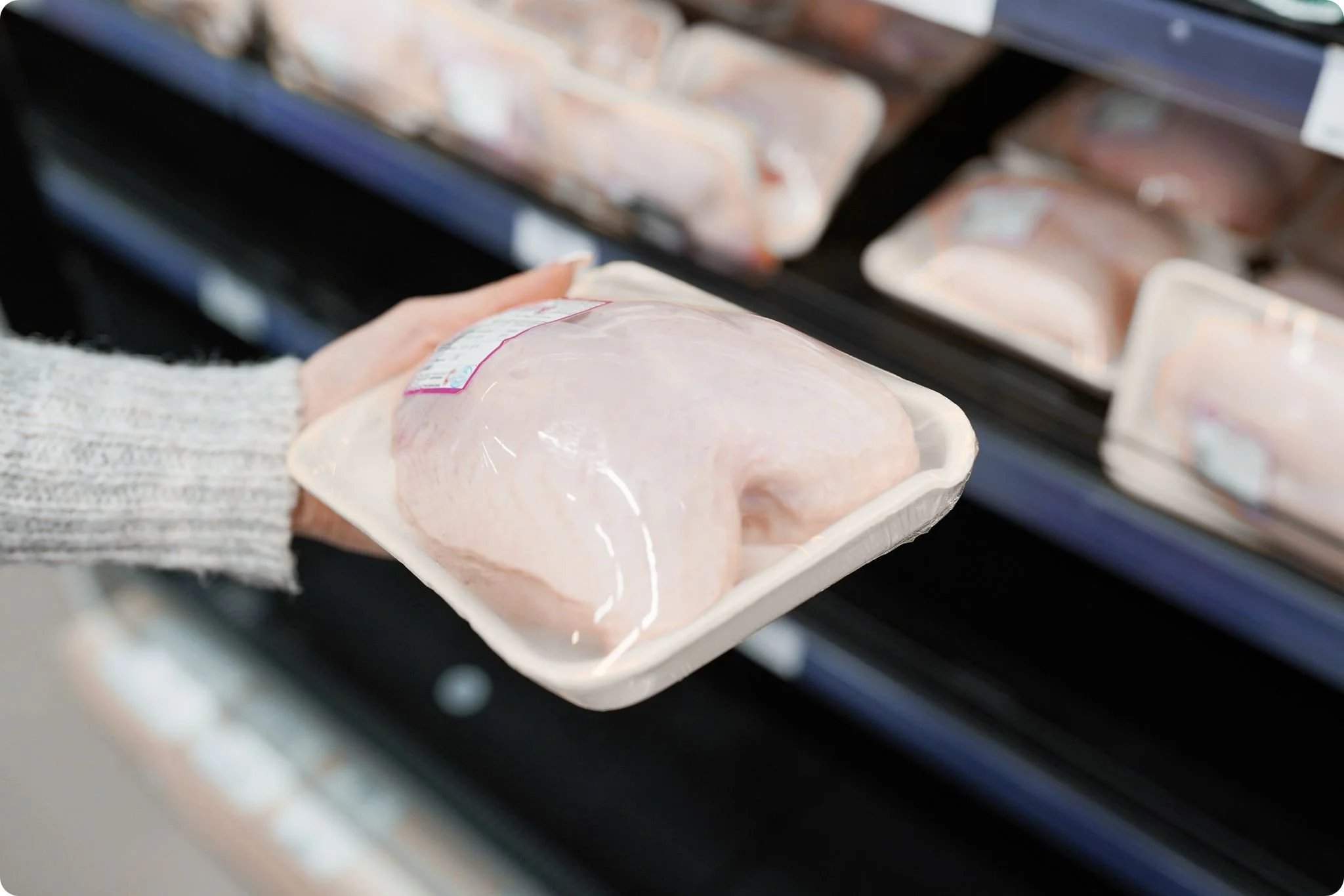 A hand wearing a gray sweater holding packaged raw chicken in a grocery store refrigerated section.