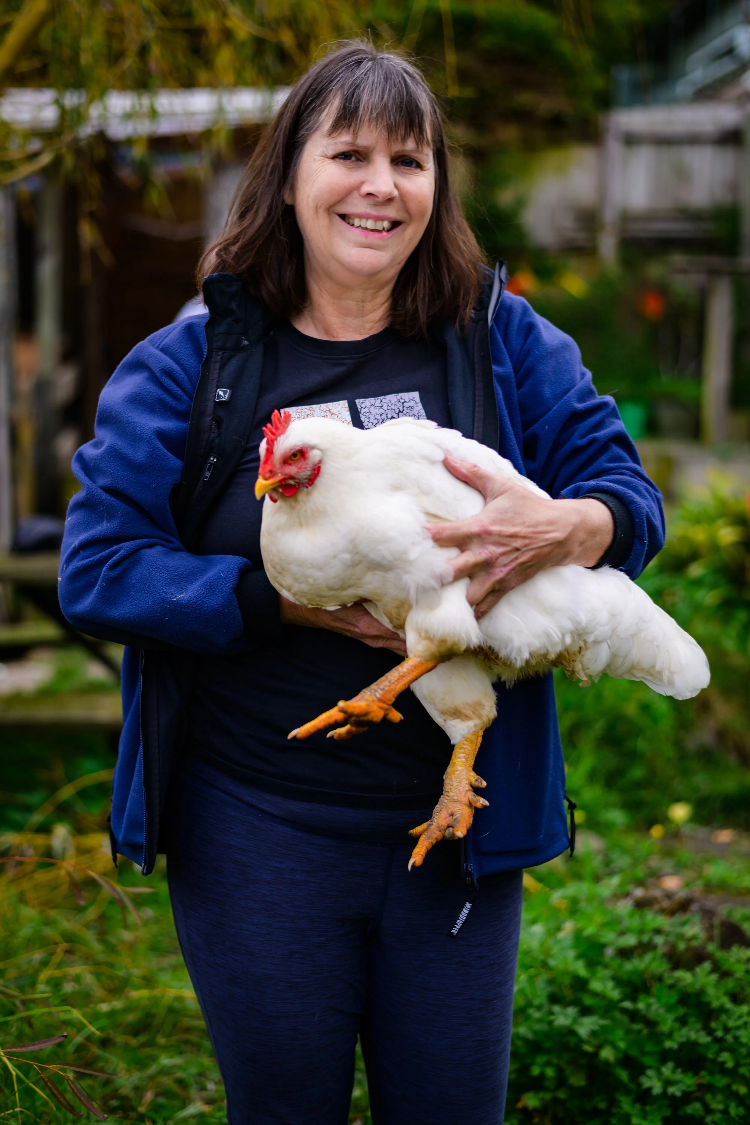 A woman with dark hair in a blue jacket holding a white chicken outdoors in a garden. The chicken is Barnaby, a rescued fast-growing meat chicken. He requires specialist care due to his extreme growth.