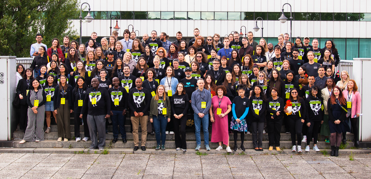 A large group of diverse people gathered outside in front of a white modern building, posing for a group photo at the Open Wing Alliance Global Summit in 2025. Many are wearing black shirts and yellow lanyards with animal advocacy messages.