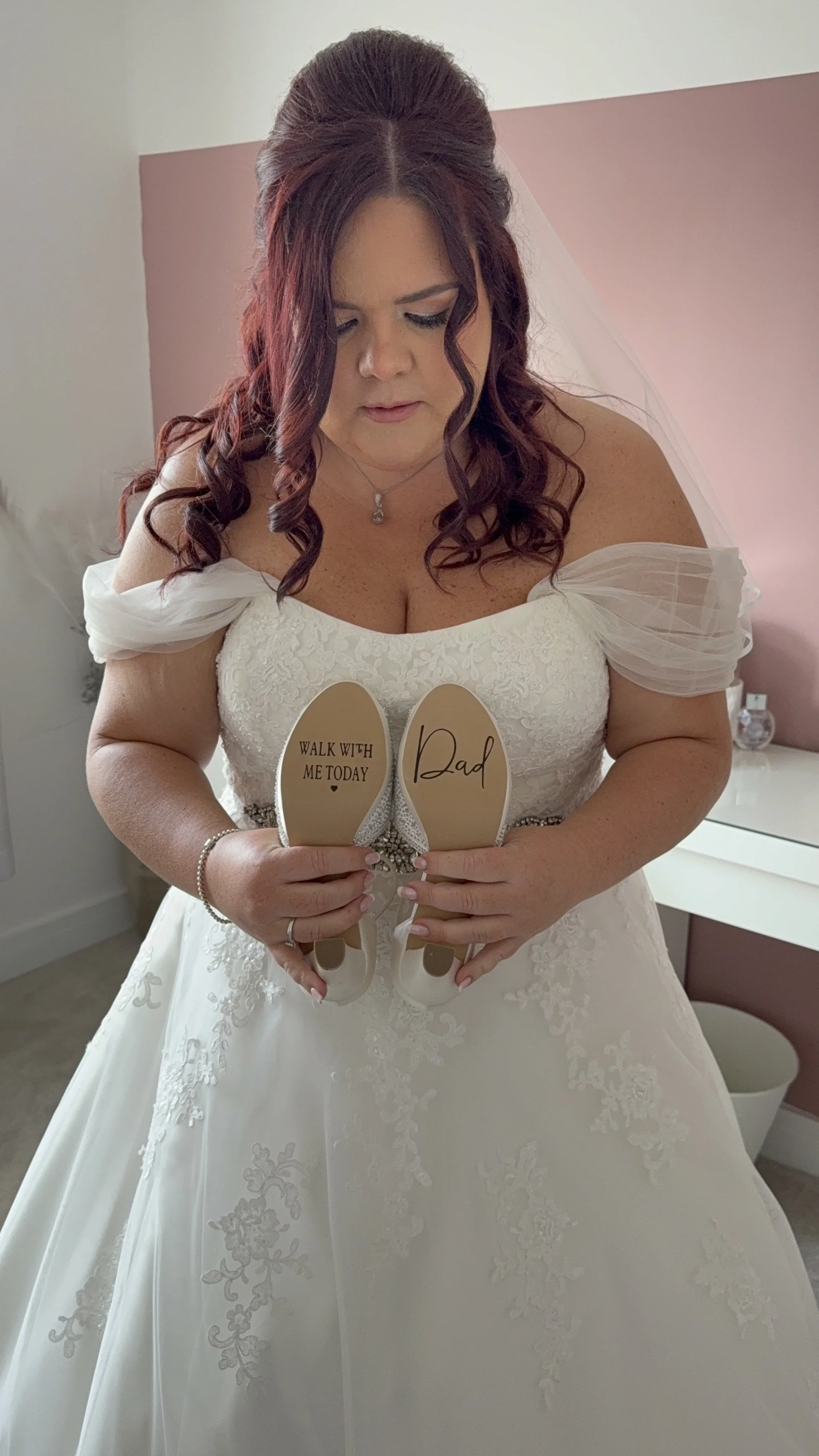 Woman in a wedding dress holding a pair of shoes, one with the message 'Walk with me today' and the other with the message 'Dad.'