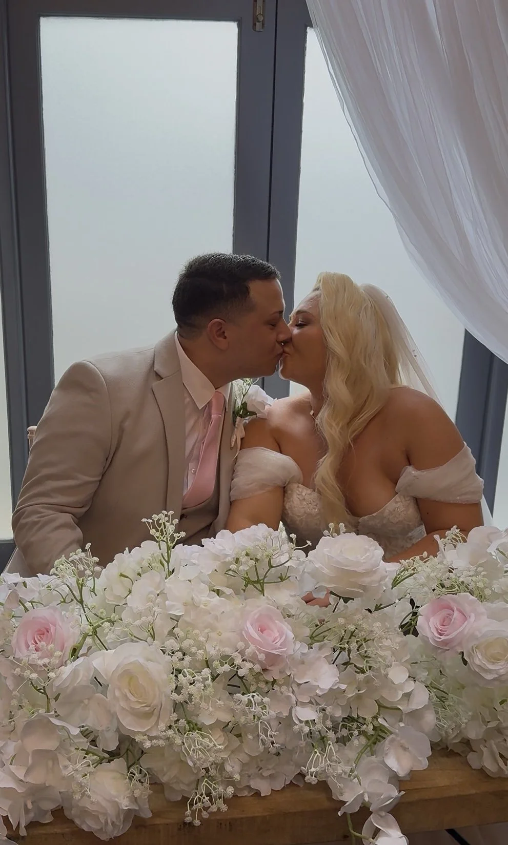 A bride and groom sharing a kiss at their wedding ceremony, sitting behind a floral arrangement of white and pink roses in a church or venue with large windows and curtains.