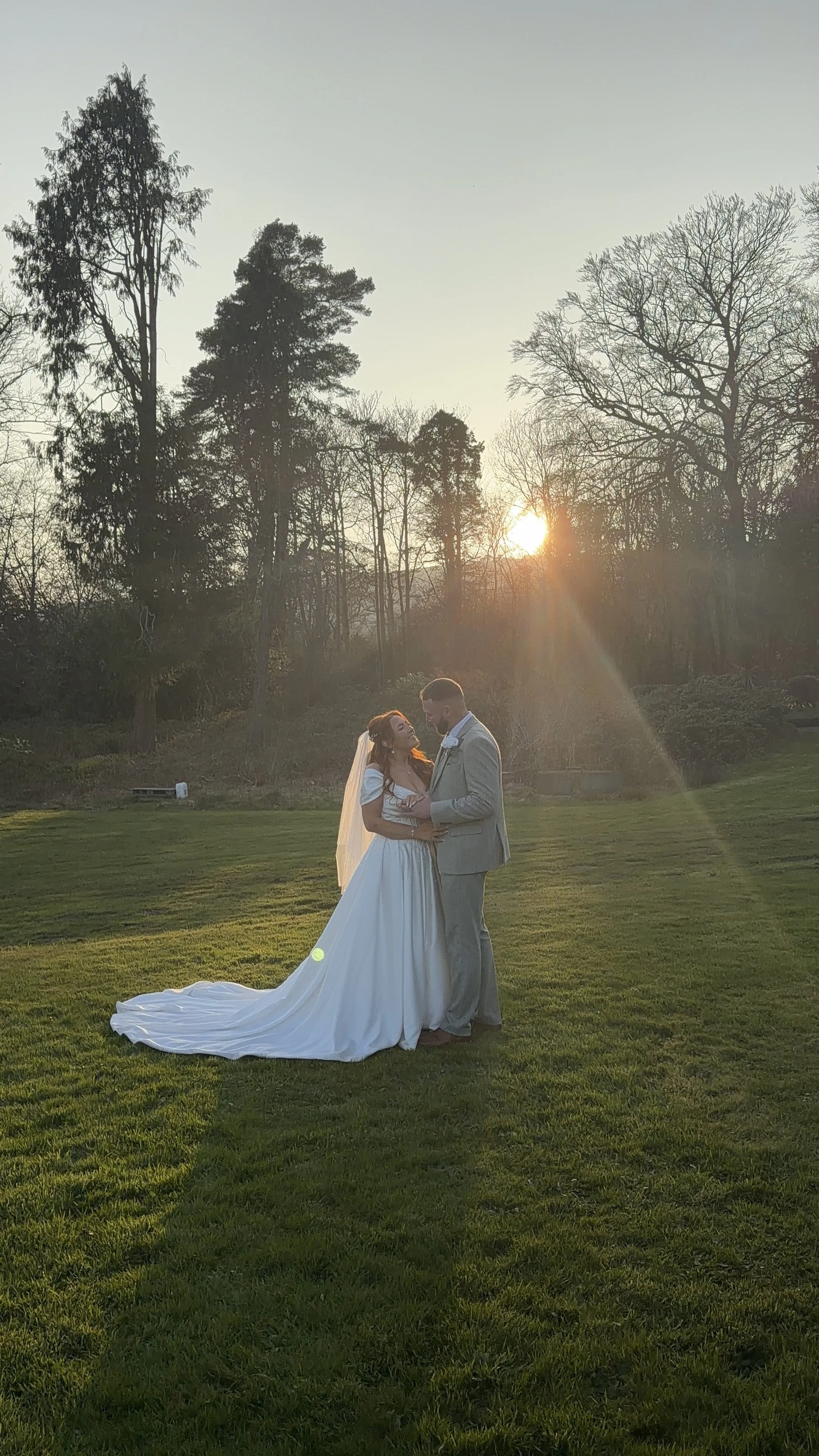 A bride and groom standing together on a grassy field during sunset, with trees in the background, embracing and looking at each other lovingly.