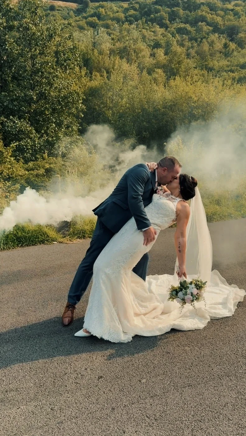 A bride and groom sharing a kiss on a rural road, with trees and smoke in the background, during their wedding photoshoot.