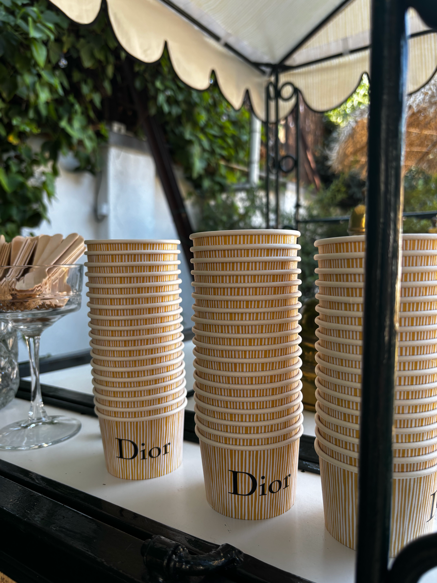 Vasos de papel con rayas amarillas y blancas, con la marca Dior escrita en ellos, apilados en una mesa al aire libre.
