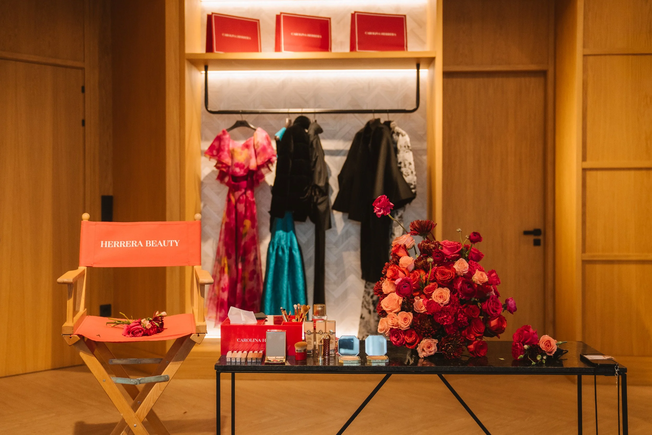 Mesa con productos de belleza y maquillaje, flores rosas y un asiento de director con la marca Carolina Herrera, en un ambiente bien iluminado con paredes de madera.