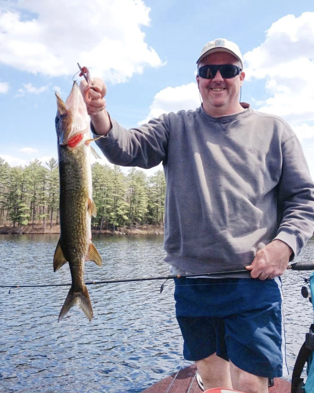 Man wearing sunglasses and a hat holding a large fish he caught while fishing at a lake with trees in the background.