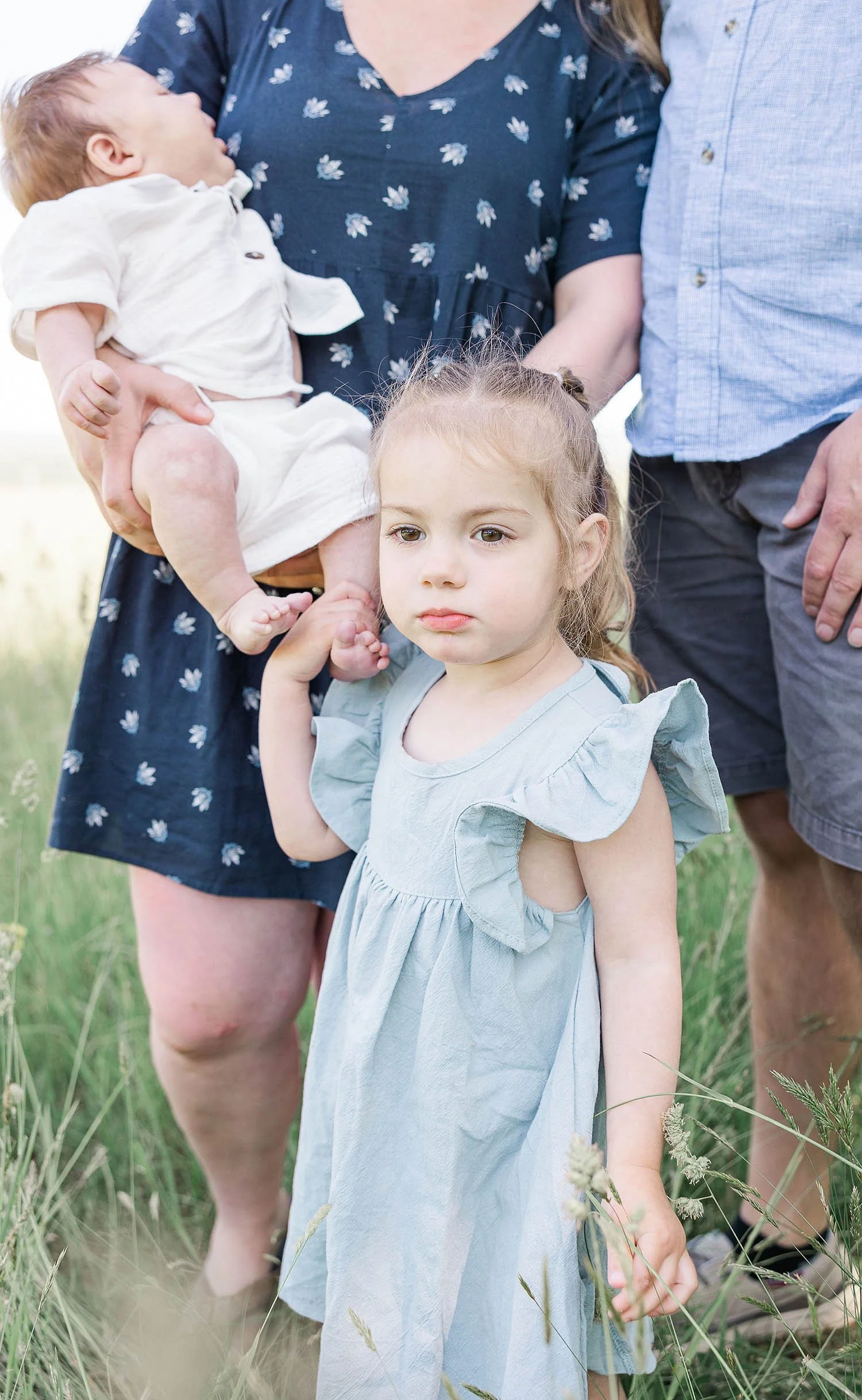 Young girl in a light blue dress standing in tall grass, with a serious expression on her face, holding someone's hand.