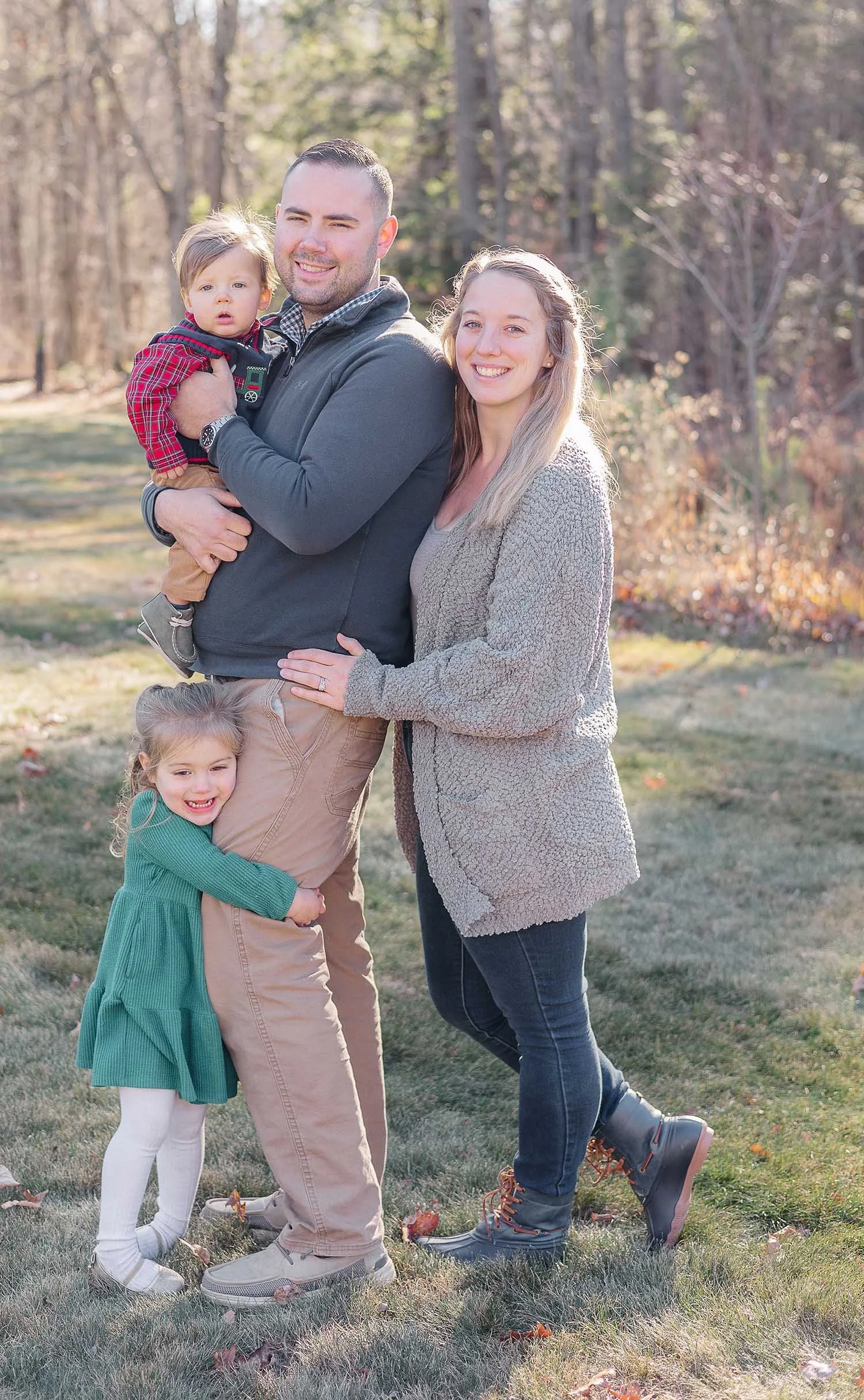 A family of four standing outdoors in a park during autumn, smiling at the camera. The father is holding a young boy, and a little girl is hugging the father's leg. The mother stands next to them, smiling, with trees in the background.