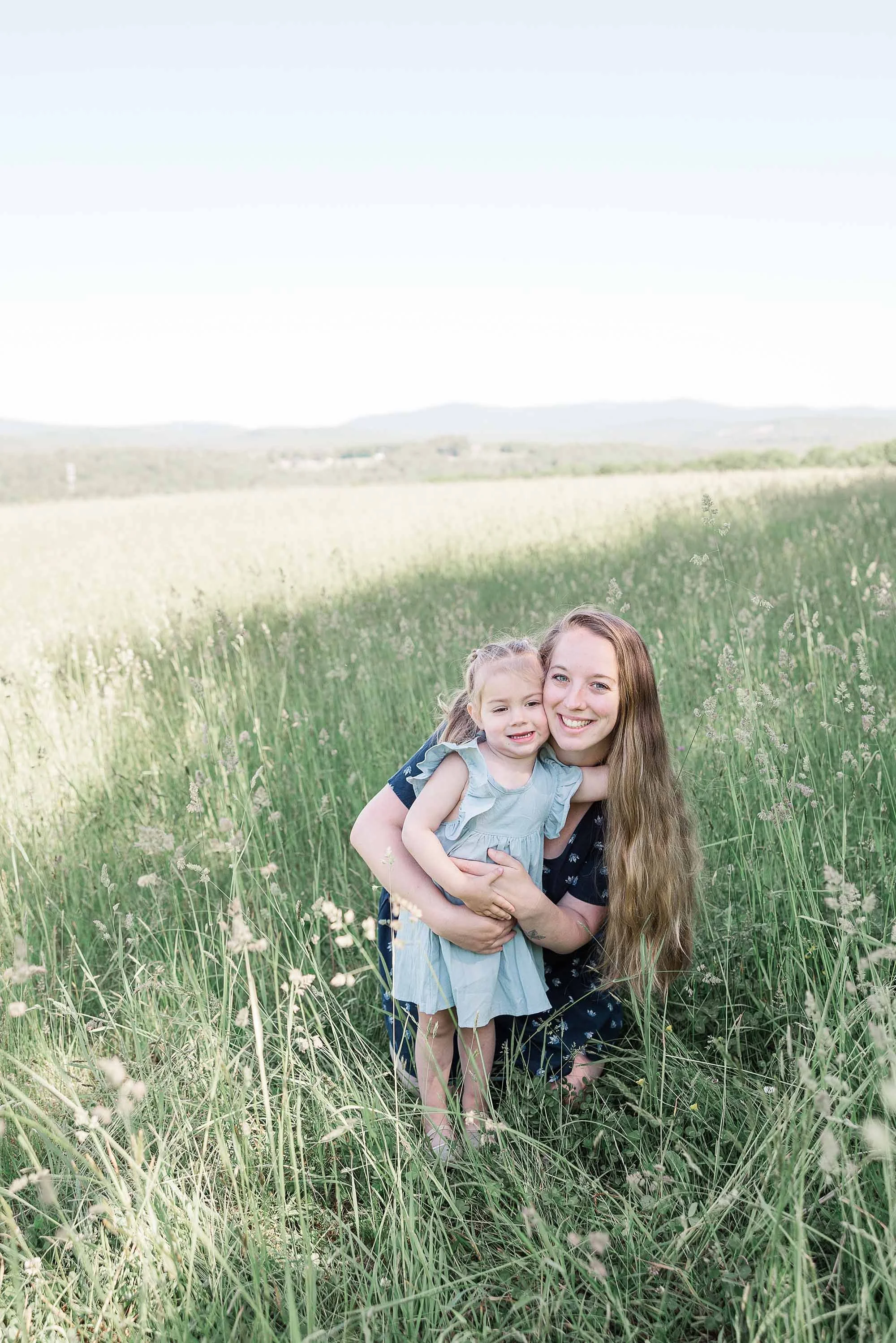 A woman with long, light brown hair and a young girl with blonde hair and a light blue dress, smiling and hugging, in a green field with tall grass, under a clear sky.