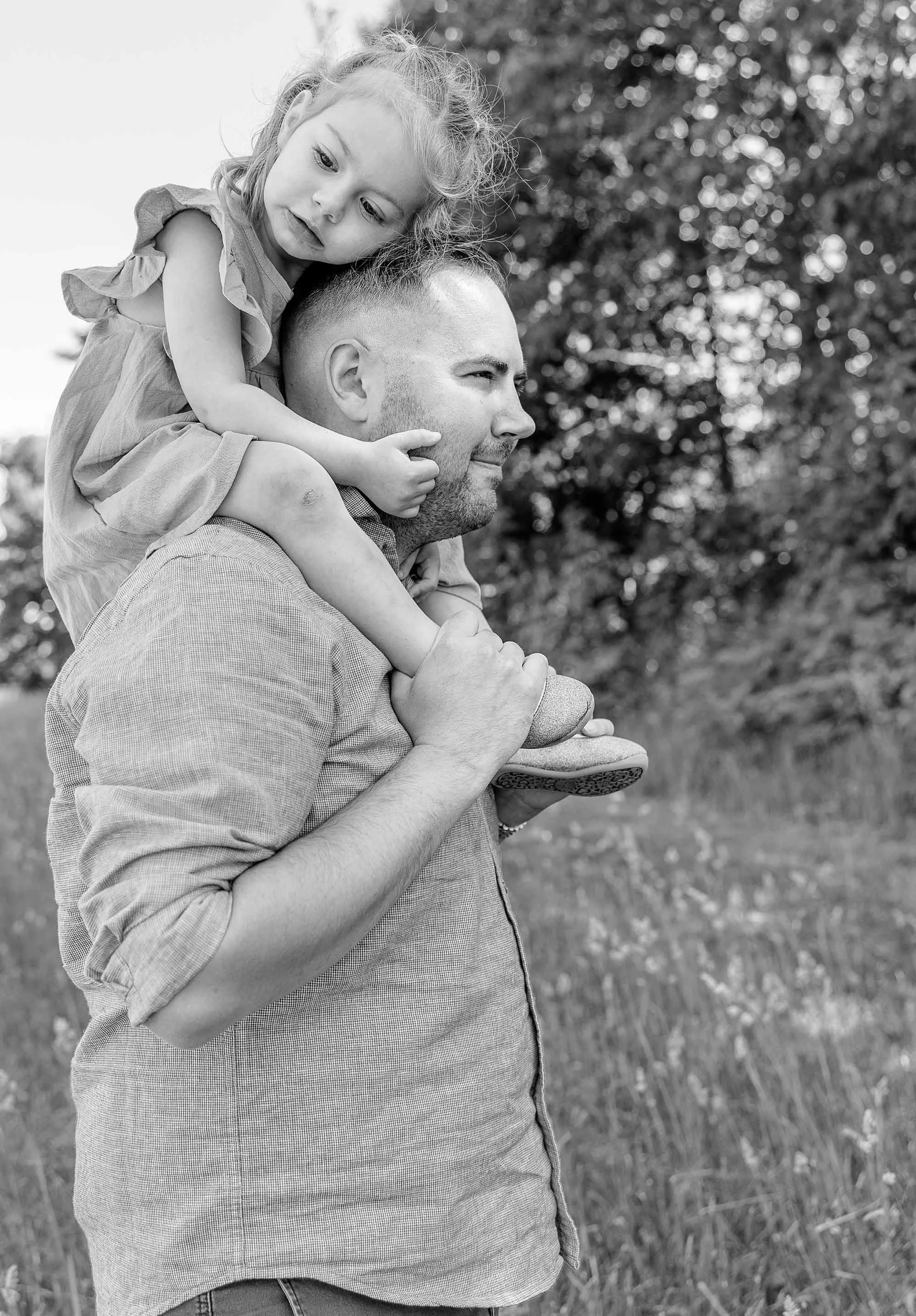 A man is carrying a young girl on his shoulders outdoors, with trees and grass in the background. The girl is resting her head against the man's head and looking down, while he smiles gently and looks ahead.